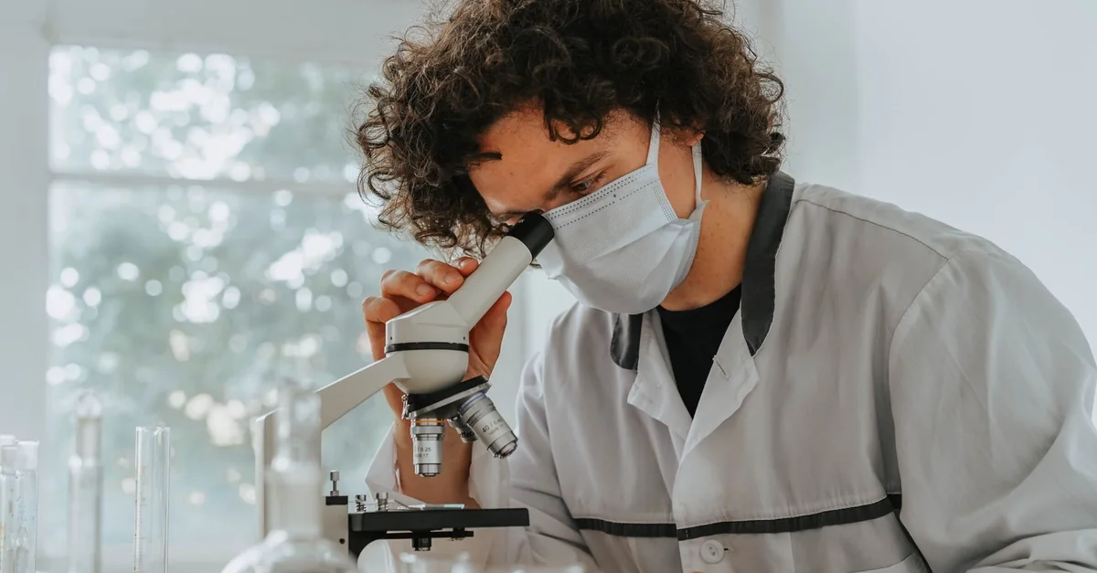 A scientist wearing a face mask examines samples under a microscope in a laboratory setting.